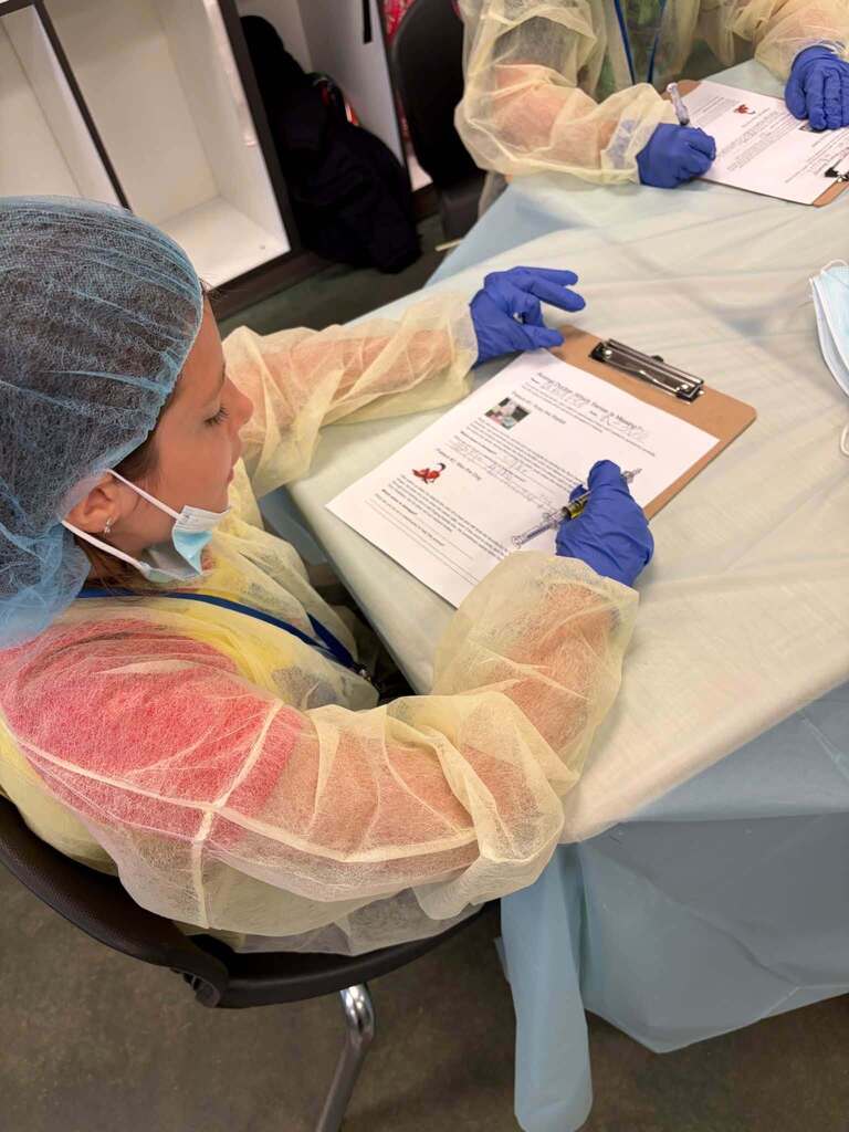A child wearing a hair net and surgical gown sits at a table in a classroom and writes on a piece of paper.