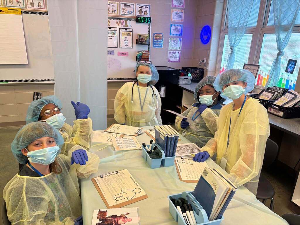 Students wearing hair nets and surgical gowns write on pieces of paper while sitting at tables.