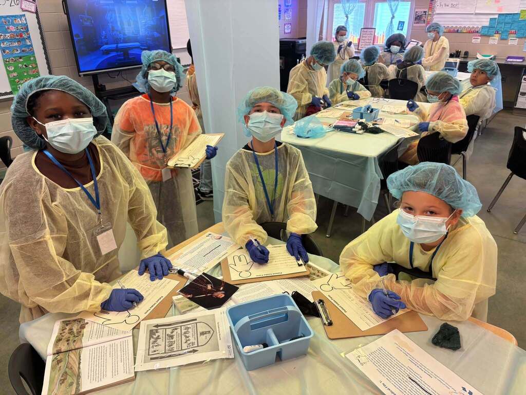 Students wearing surgical gowns, hair nets, and masks write on pieces of paper while sitting and standing at desks in a classroom.