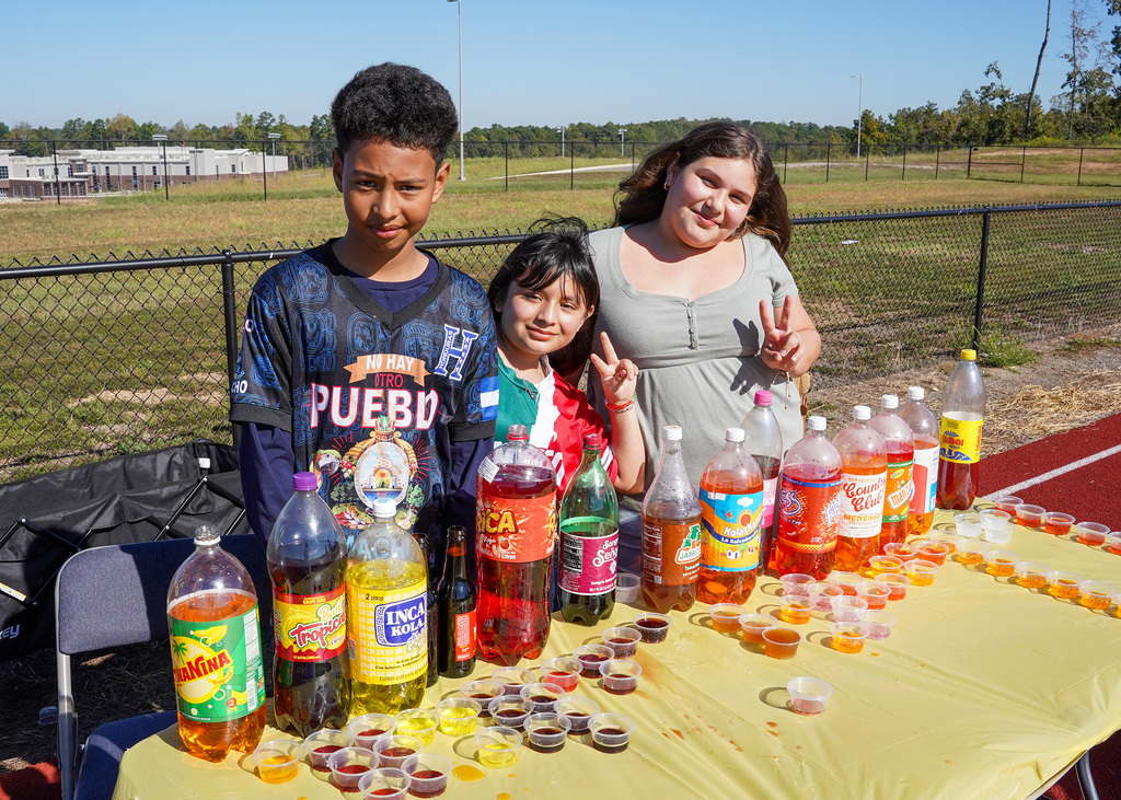 Students smile while standing behind bottles of soda set up on a table outdoors.
