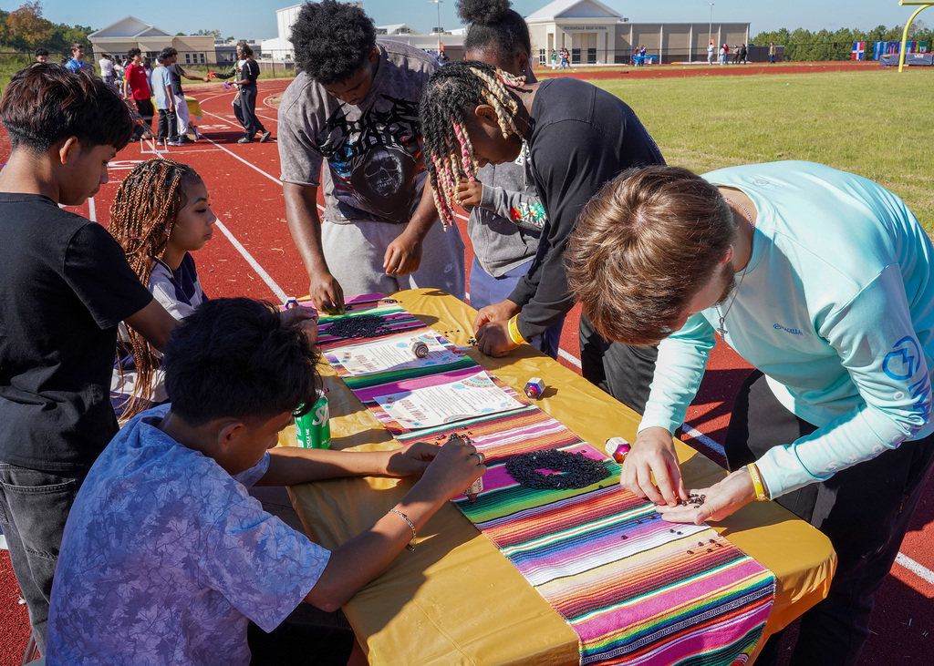 Students sit and stand at a table set up on a track outdoors and play a board game.