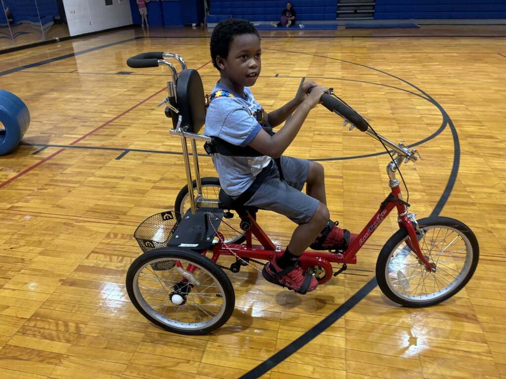 A Burkett Center student rides an accessible tricycle through a school gym.
