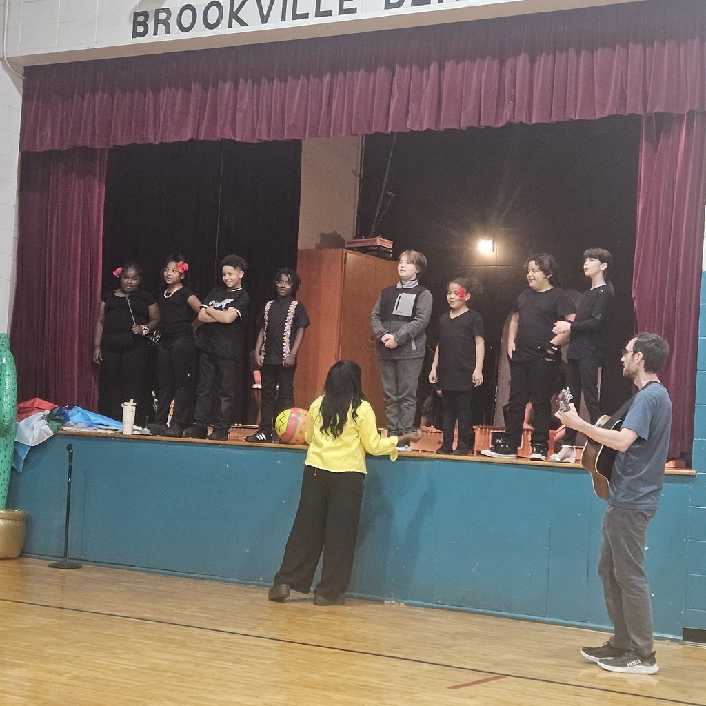 Students singing on stage while Ms. Crawford directs from the floor. Guitarist is standing on the floor playing. 