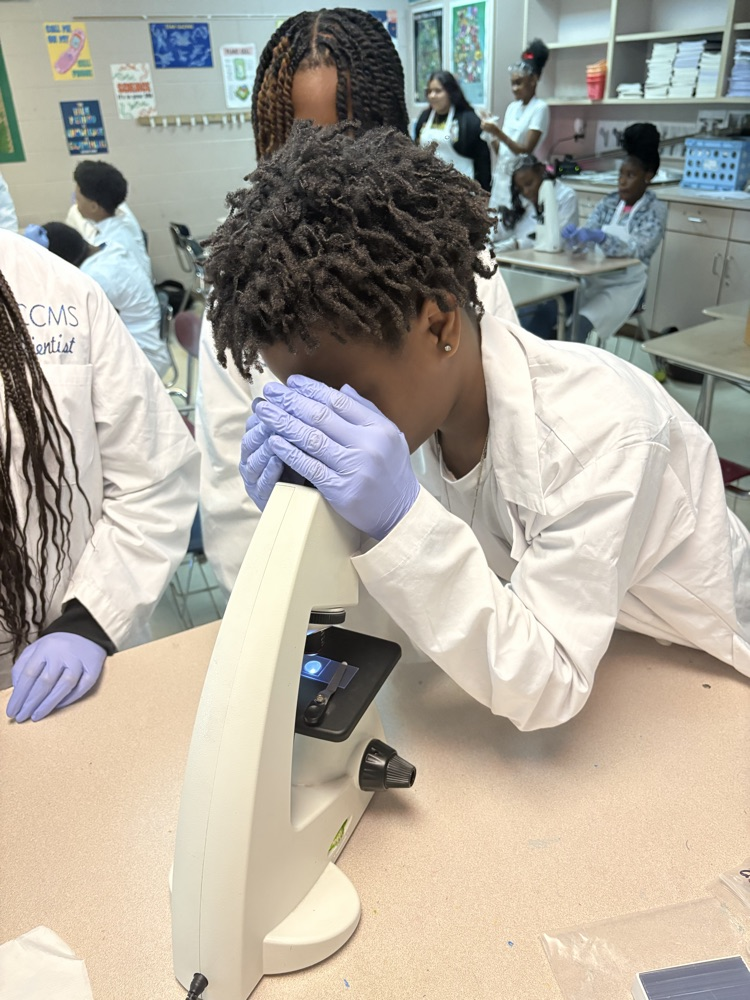 student in white lab coat and gloves looking in microscope