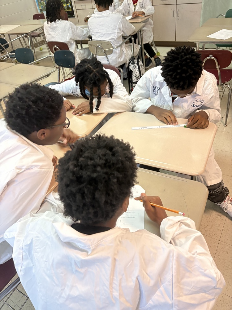 students sitting in brown desks wearing white coats and writting