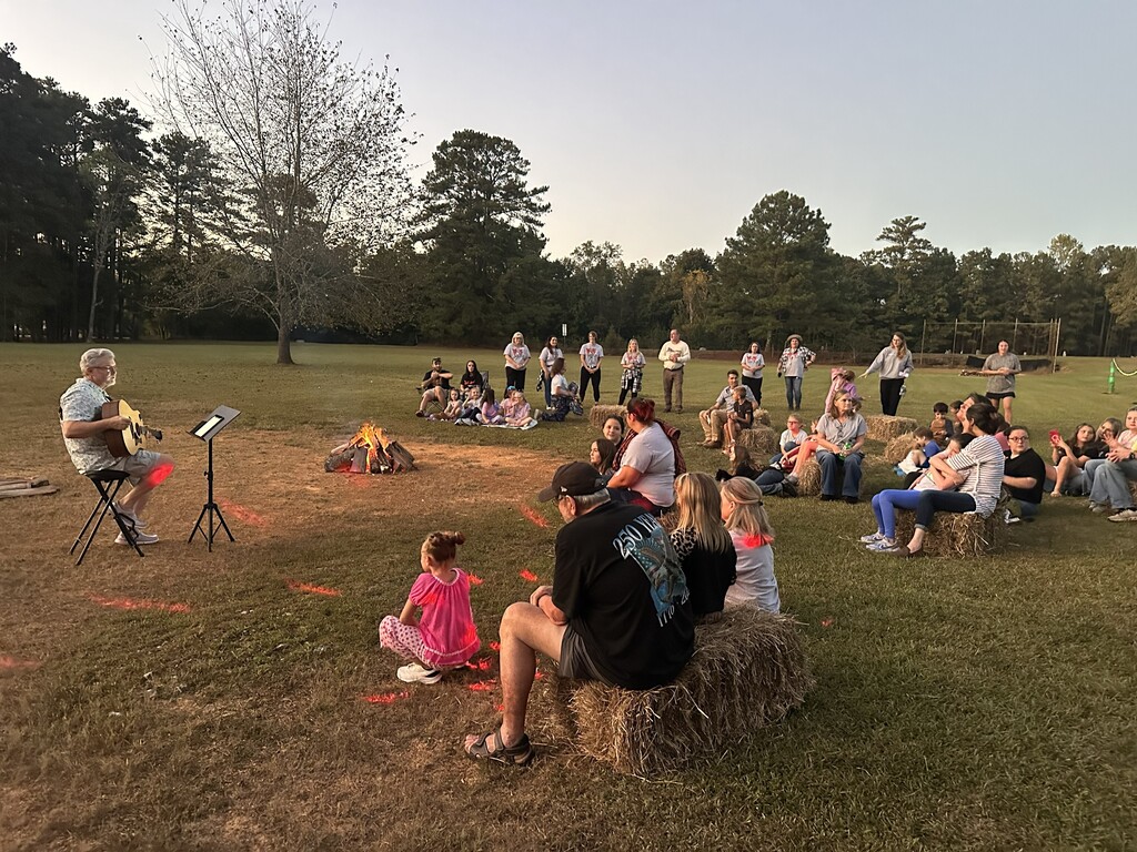 a man playing guitar around a campfire with people standing and sitting around