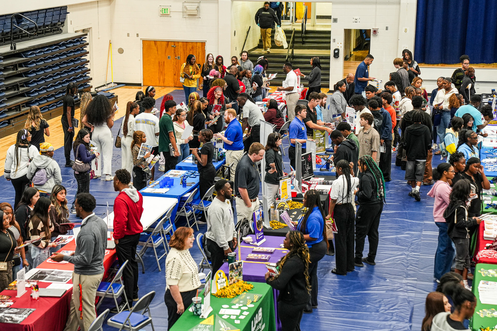 Students attend a college fair inside a school gym. College representatives stand behind tables and speak to the students.