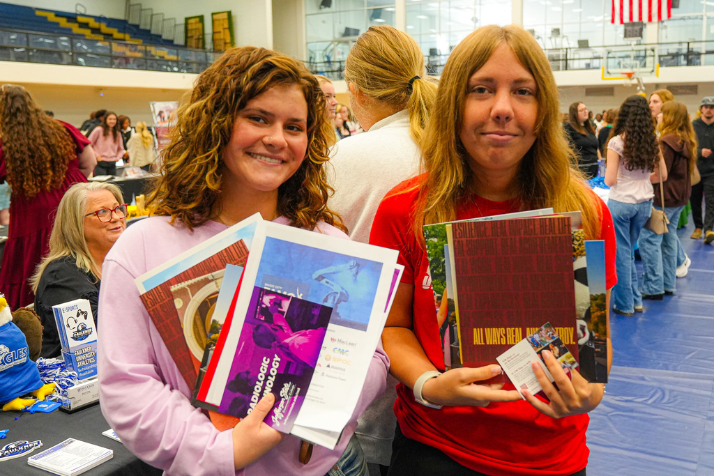 Two students attending a college fair in a gymnasium smile for a photo while holding papers they received from colleges.