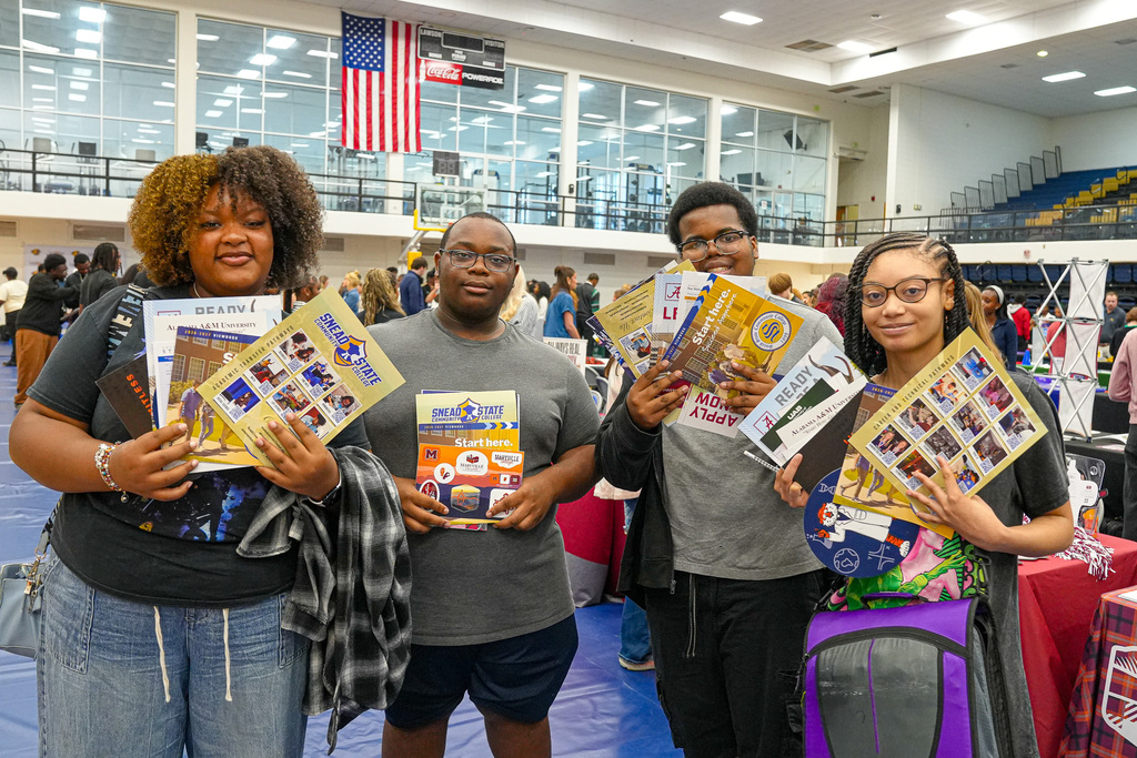 Students attending a college fair in a gymnasium smile for a photo while holding papers they received from colleges.