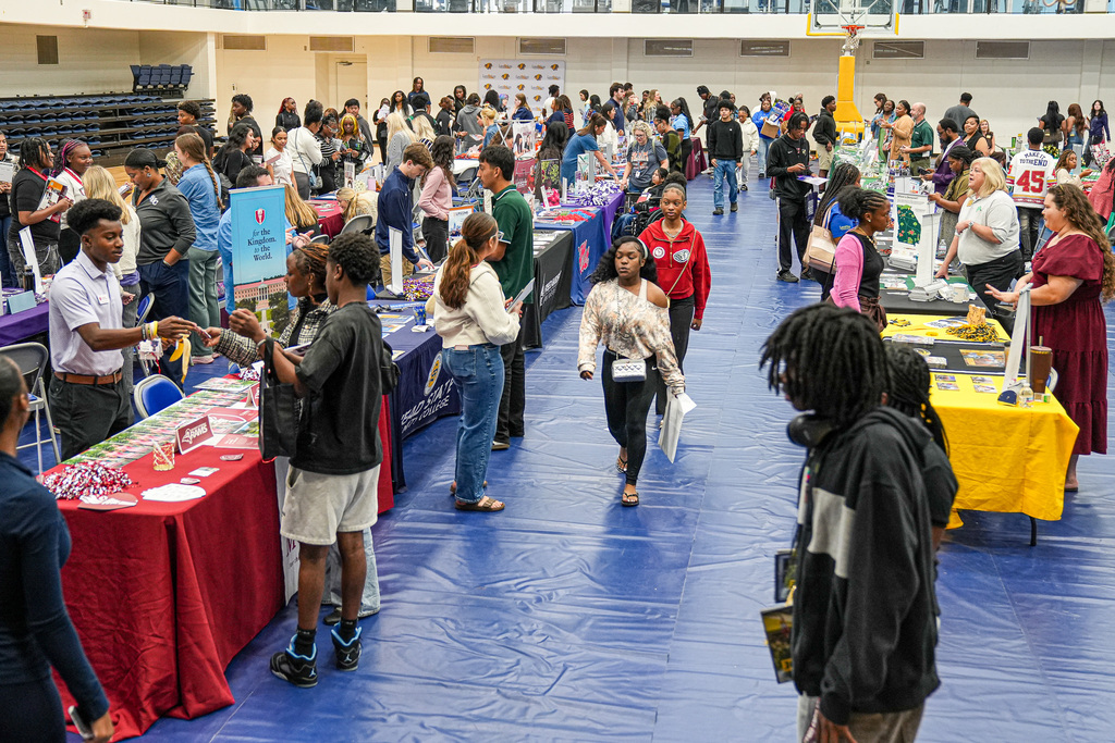 Students attend a college fair inside a school gym. College representatives stand behind tables and speak to the students.