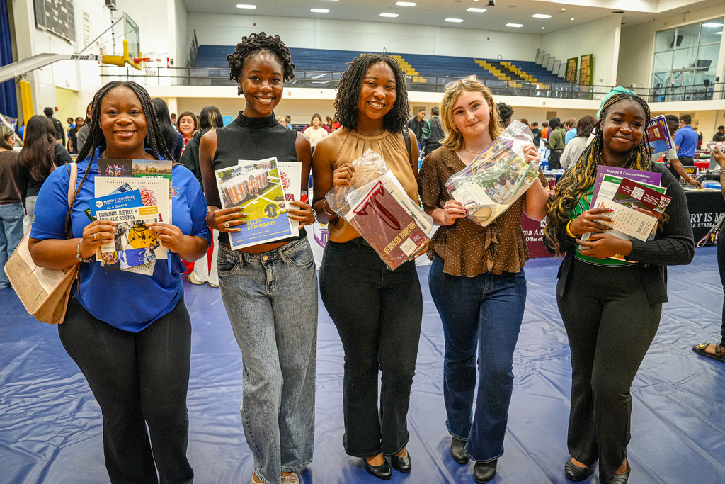 Students attending a college fair in a gymnasium smile for a photo while holding papers they received from colleges.