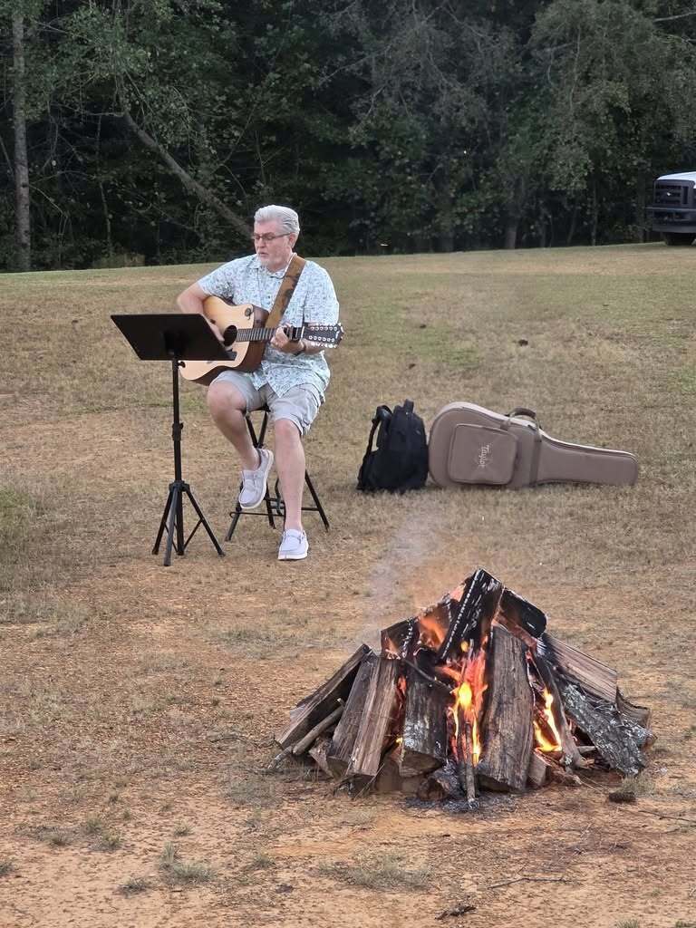 A man plays guitar while sitting in a field behind a campfire.
