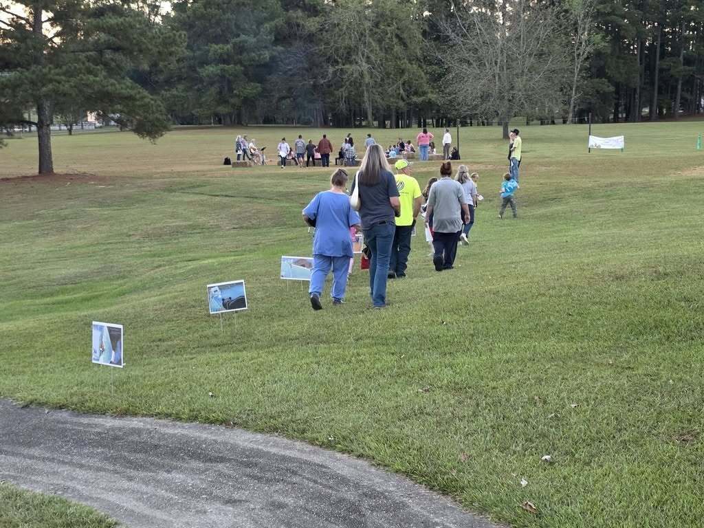 Families walk toward a crowd of people sitting around a campfire in a field.