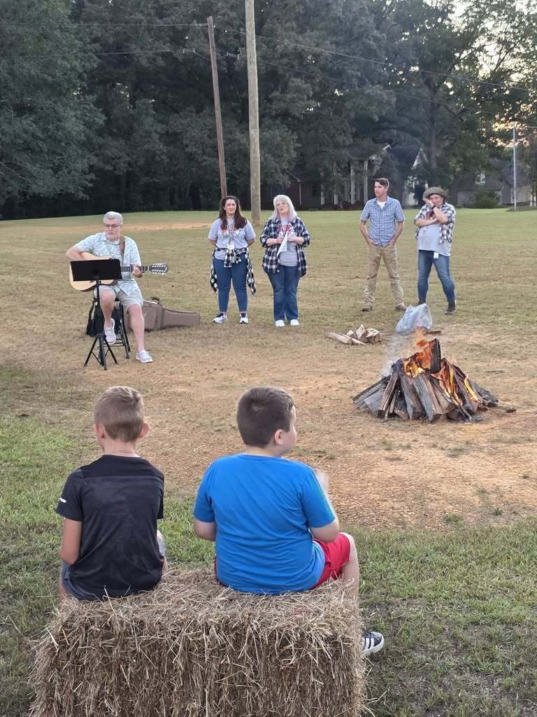 Children sit on a hay bale and listen to a man as he plays guitar in front of a campfire set up in a field.