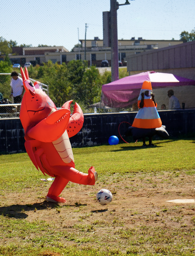 A person wearing an inflatable lobster costume kicks a soccer ball during a kickball game on a baseball field.