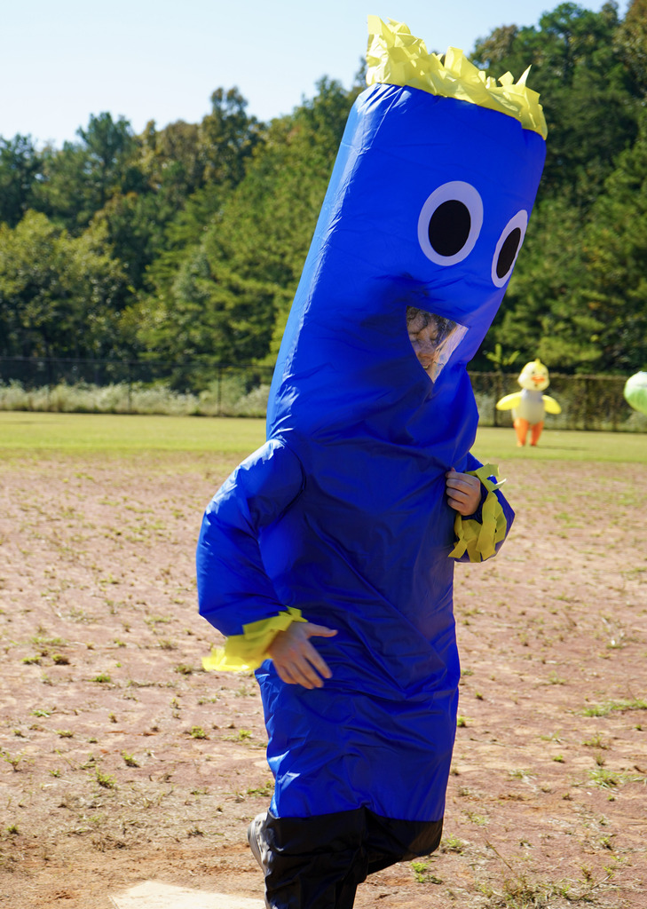 A person wearing an inflatable costume runs across home plate during a kickball game on a baseball field.