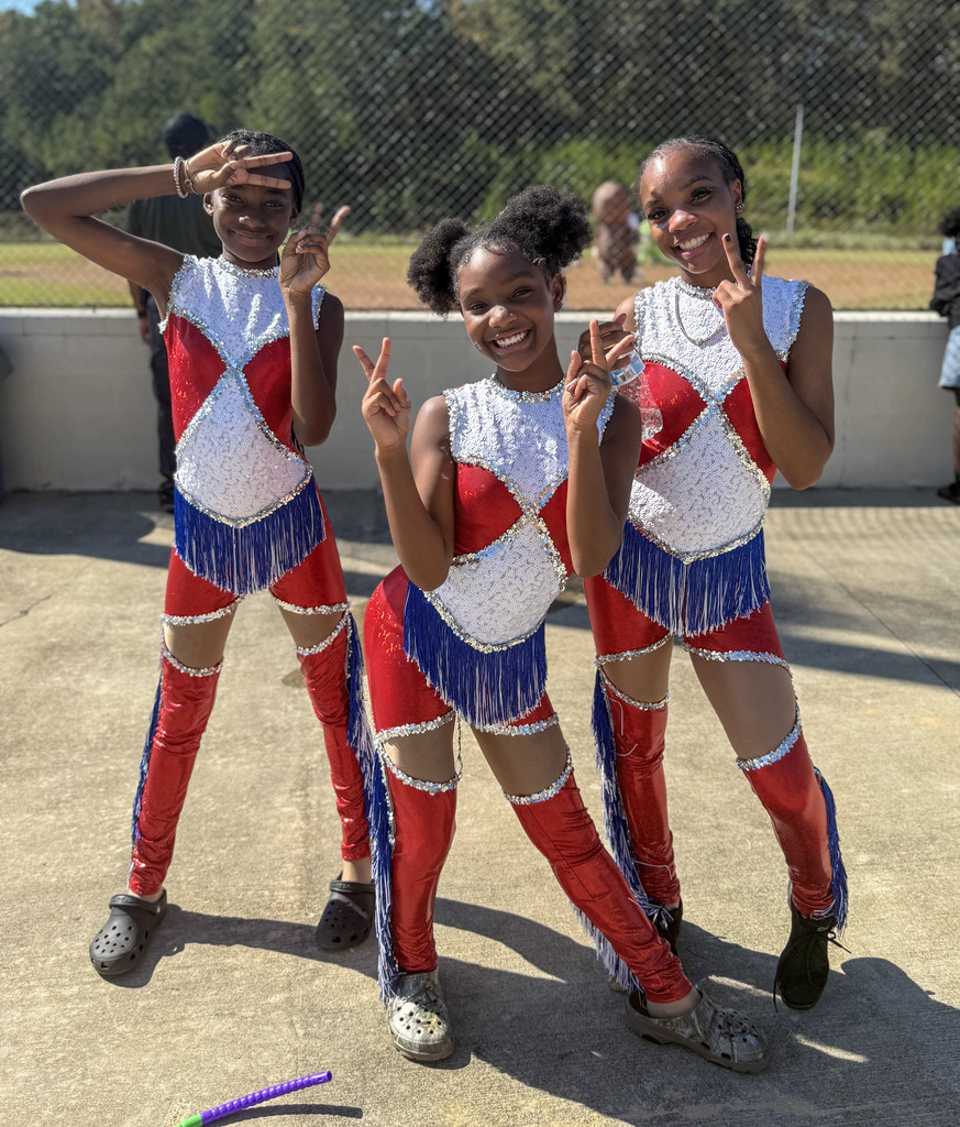 Students wearing red, blue, and white dance team costumes smile for a photo while standing outside a baseball field.