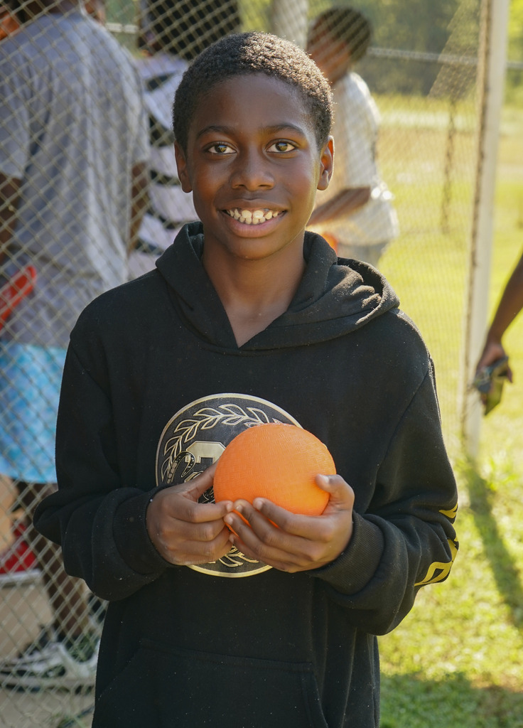 A student smiles while holding an orange ball and standing on a baseball field.