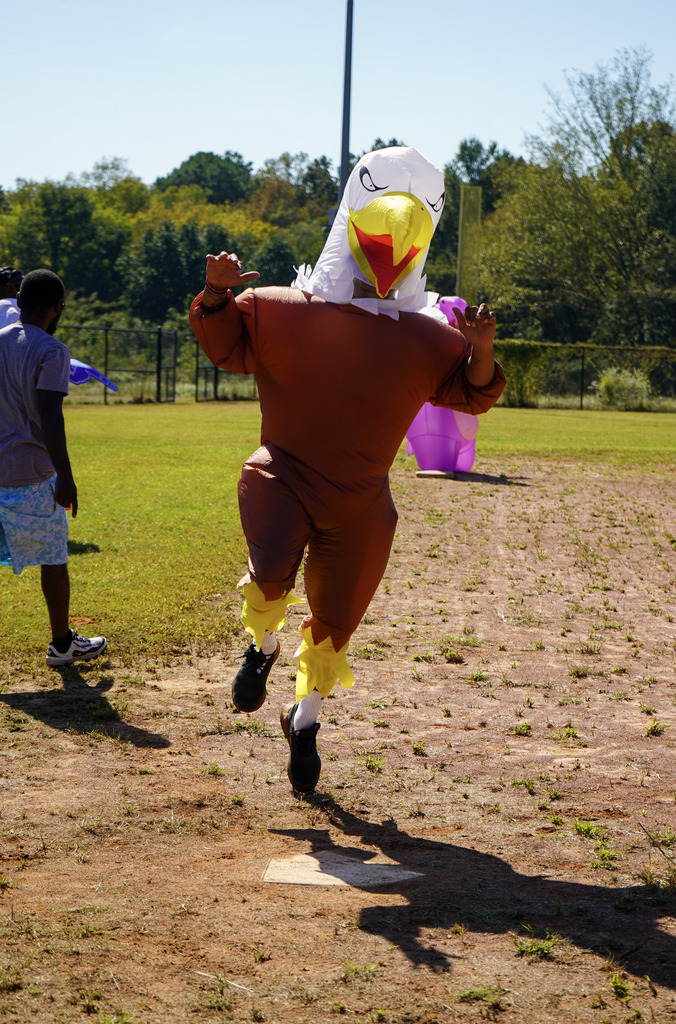 A person in an inflatable eagle costume runs across home plate during a kickball game on a baseball field.