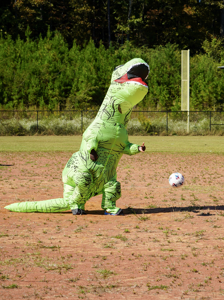 A person wearing a dinosaur costume rolls a soccer ball down a baseball field during a kickball game.