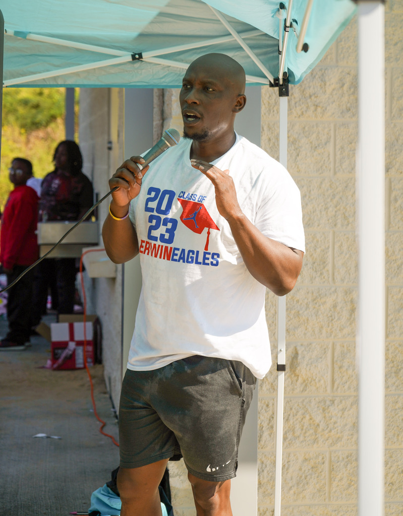 A man speaks into a microphone while standing near the concession stand of a baseball field.