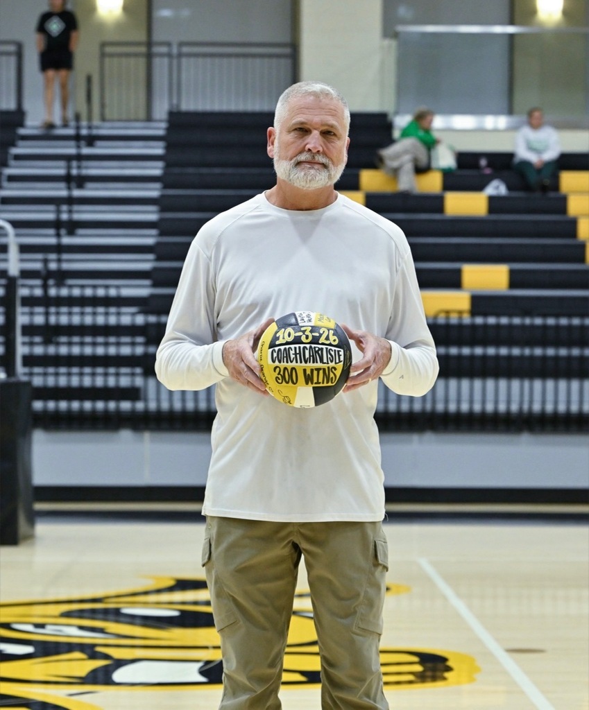 Corner High School volleyball coach Terrel Carlisle stands in a school gym and holds a volleyball in his hand. The volleyball has "10-3-26 Coach Carlisle 300 wins" written on it.
