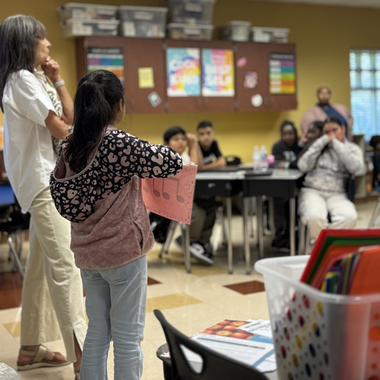 a teacher and student standing in from of a class as the student presents a music note symbol