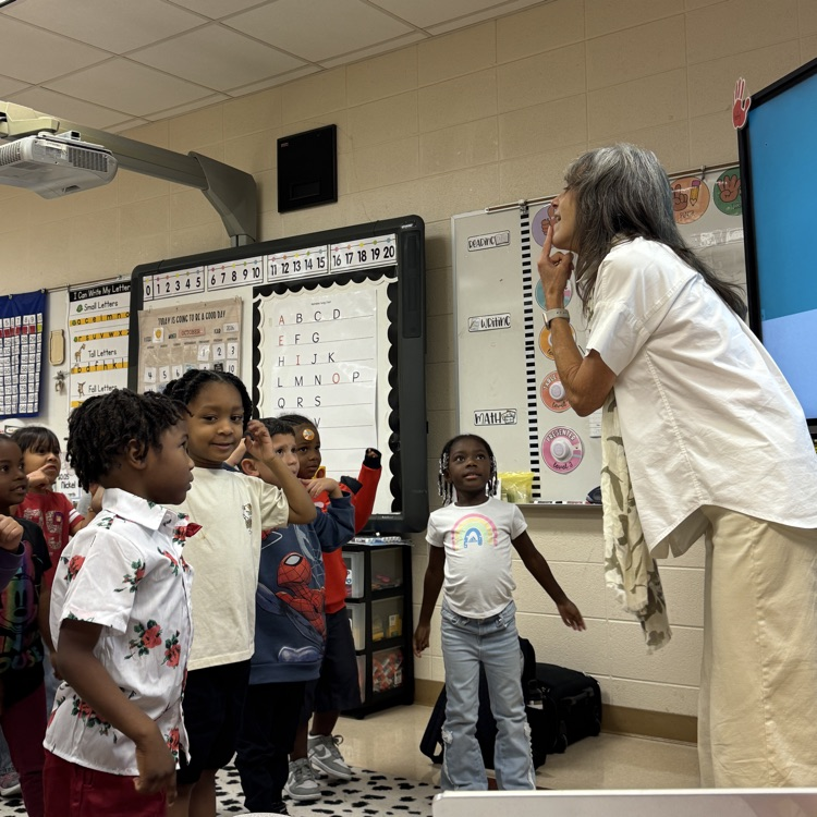 young students stand and listen to a teacher as she points to her chin