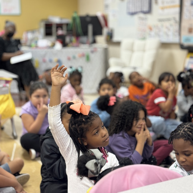 students sitting on a rug and one student has her hand raised to ask a question