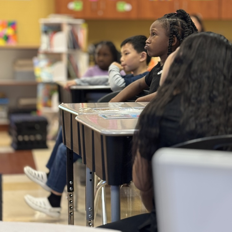 students sit at desks and listen to a teacher