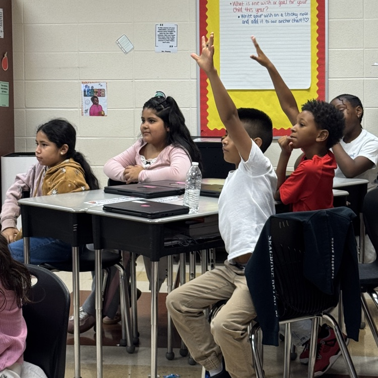 five students sitting a desks while two of the have their hands raised