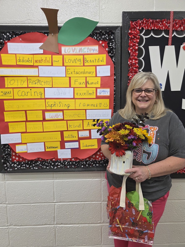 our principal standing in front of an apple with words that describe her. She is holding flowers and a gift.