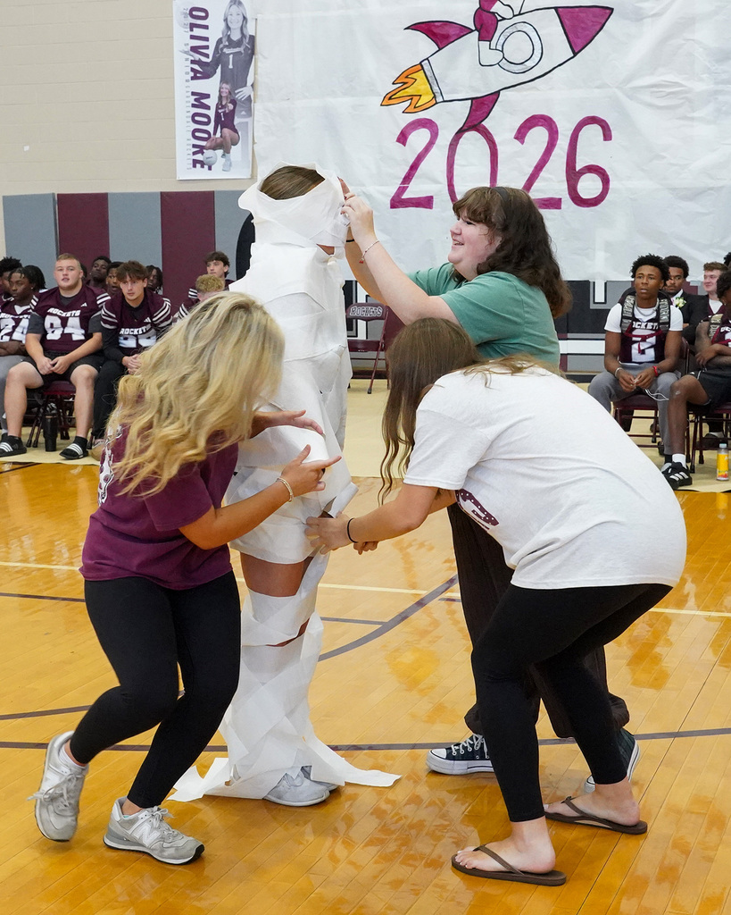 People wrap a student in toilet paper during a pep rally game in a school gym.