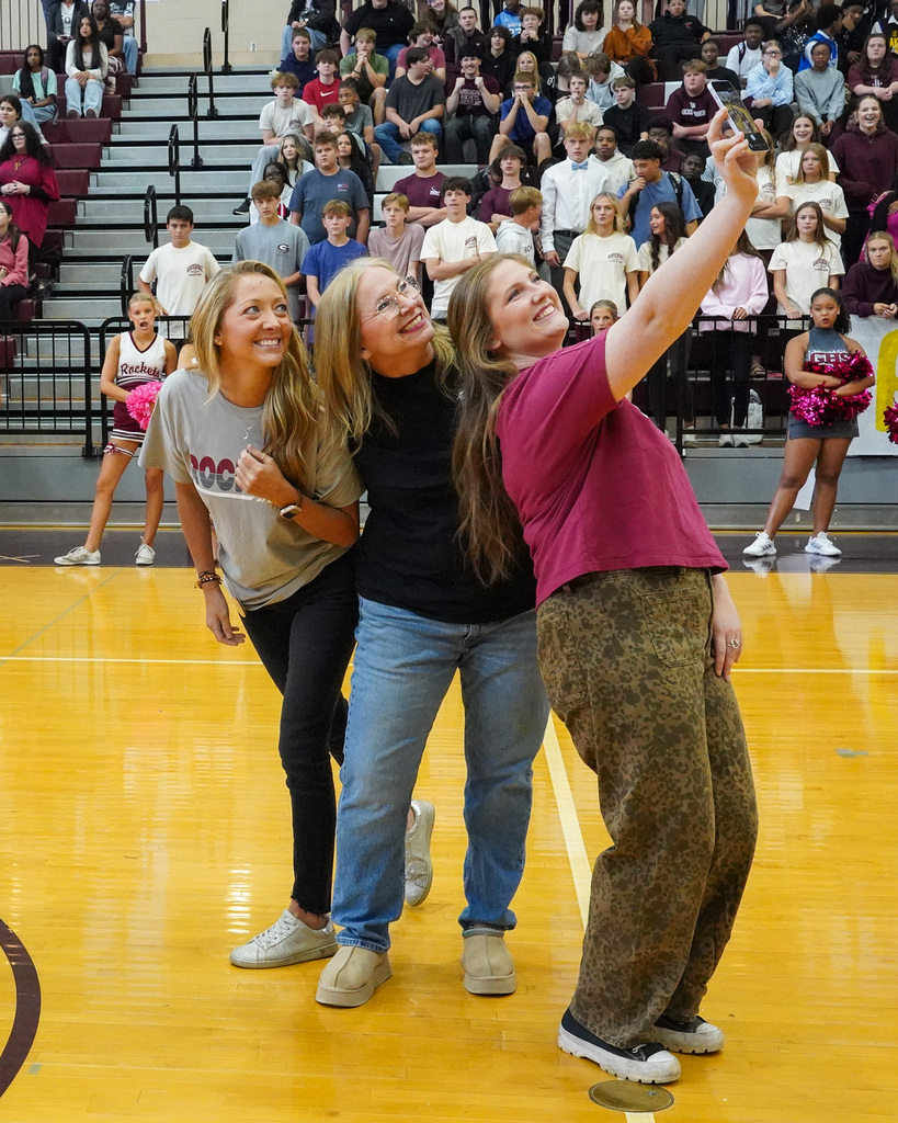 Three women take a selfie while standing on the gym floor during a pep rally.