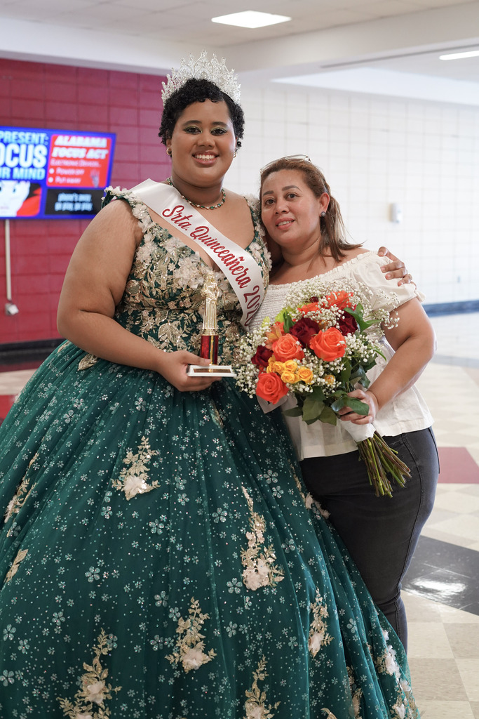 A student wearing a quinceañera gown and a crown poses for a photo with her mom in the lobby of a school auditorium.