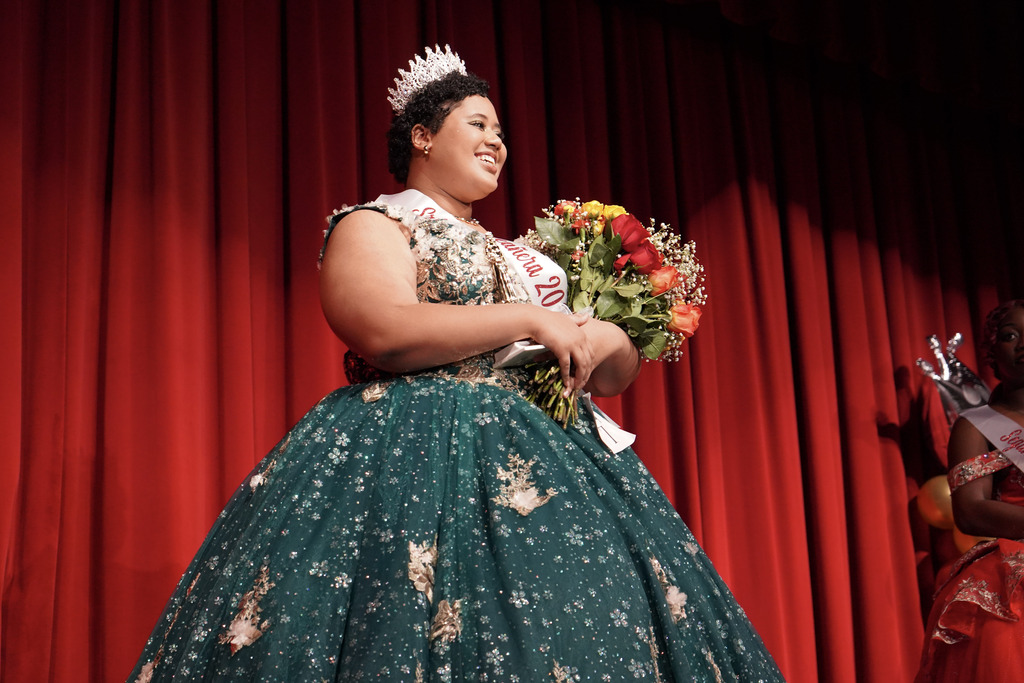 A student wearing a sparkly quinceañera gown, crown, and sash smiles for a photo while standing on a stage and holding flowers.