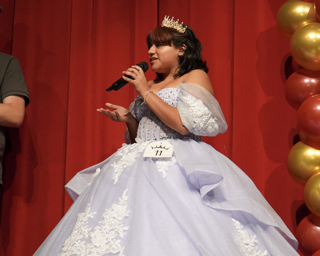 A student wearing a purple quinceañera speaks into a microphone while standing on a stage.