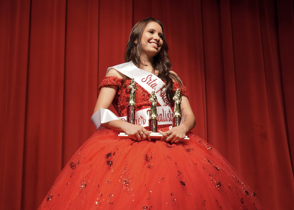 A student wearing a quinceañera gown and sashes holds trophies and smiles while standing on a stage.