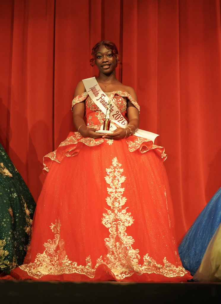 A student wearing a red ballgown smiles for a photo while standing on a stage. She wears sashes and holds a trophy.
