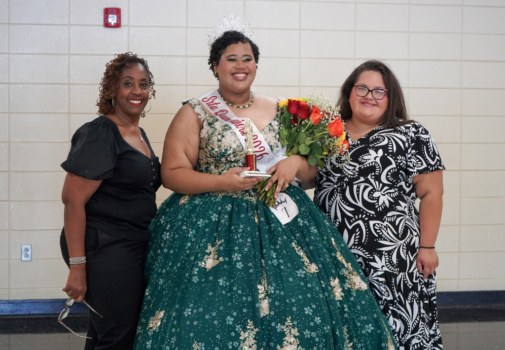 A student in a quinceañera gown and a crown smiles for a photo with two women.