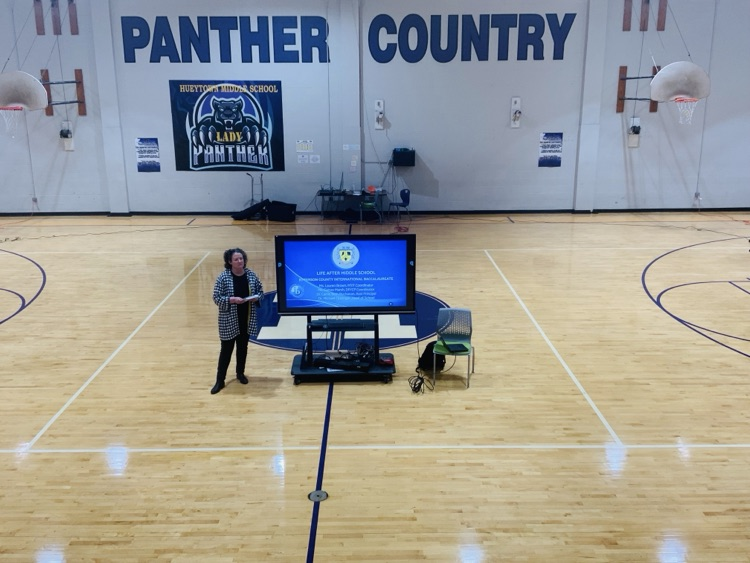 A presenter stands beside a screen displaying JCIB recruitment information in the HMS competition gym.