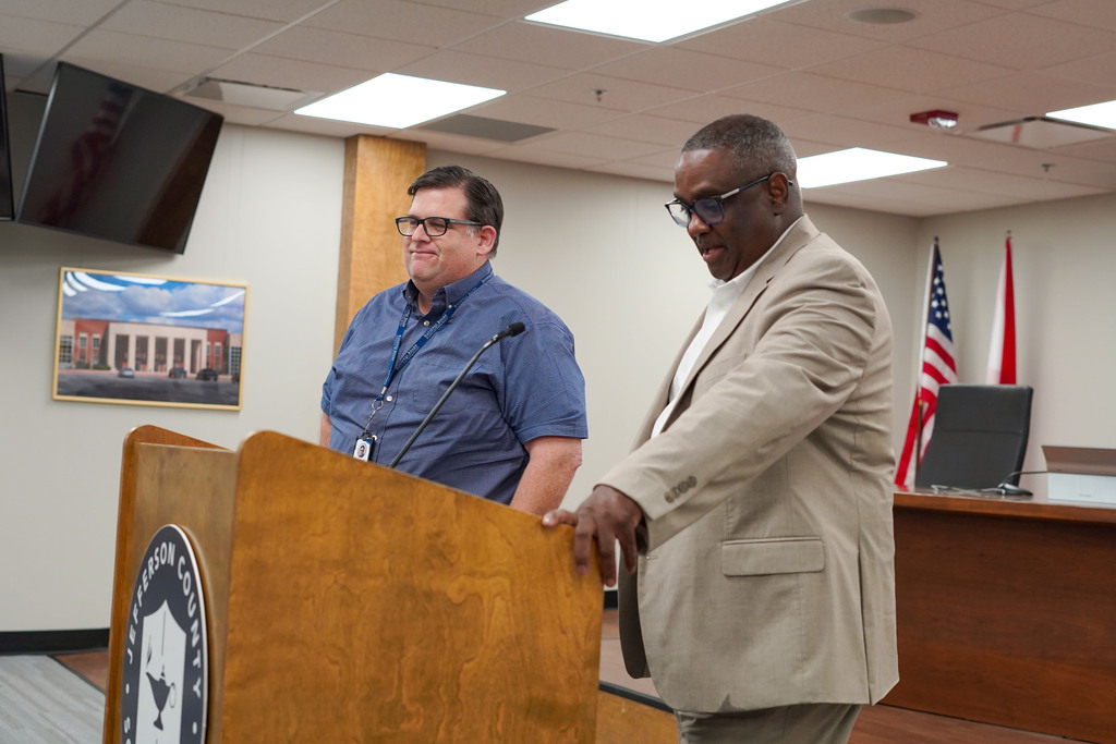 Paul Kelsey stands next to Superintendent Dr. Walter Gonsoulin in the JEFCOED Board Room. Dr. Gonsoulin speaks into a microphone behind a podium.