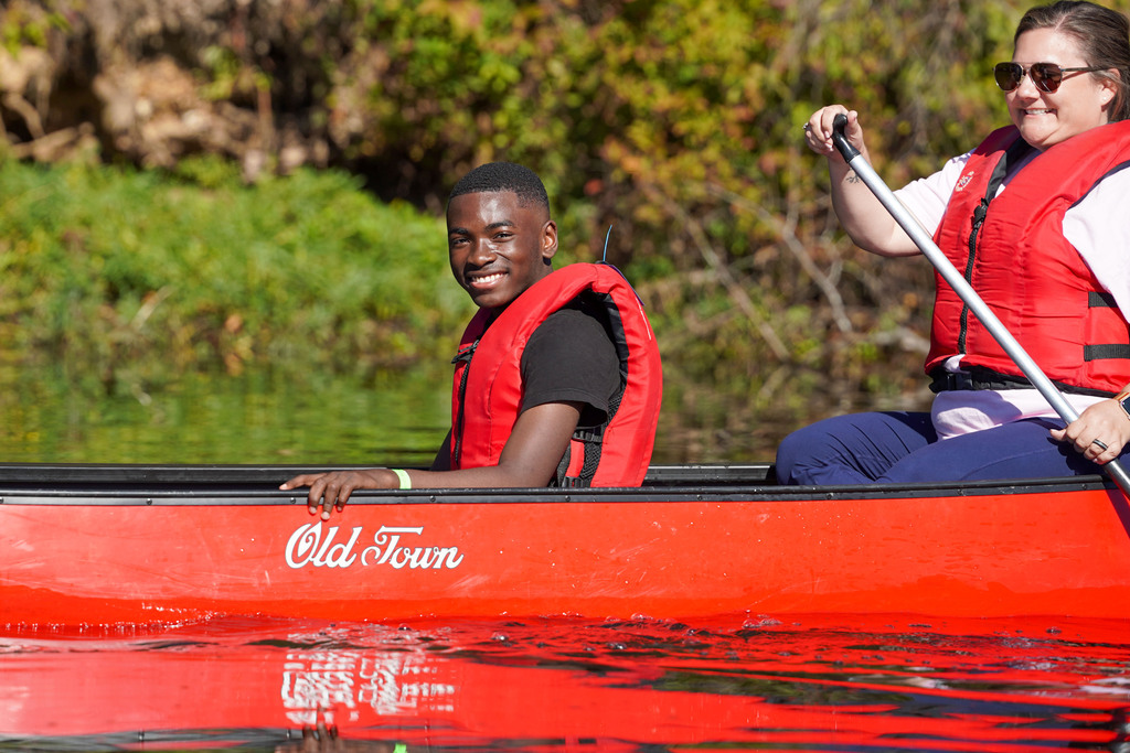 A student smiles while sitting in the middle of a canoe being paddled by two women.