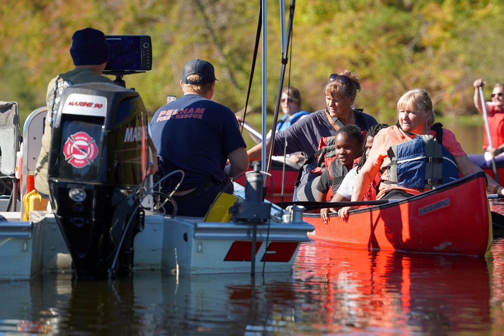 Pelham firefighters sitting in a boat speak to children and women as they sit in a canoe on a lake.