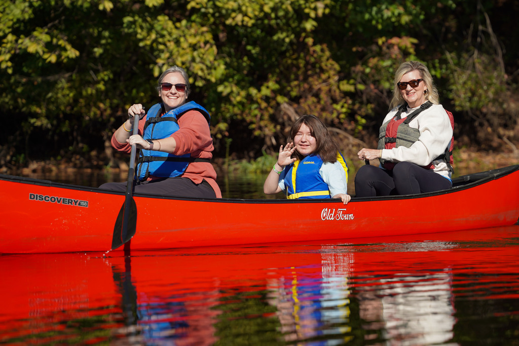 A child waves while sitting in a canoe being paddled by two women.