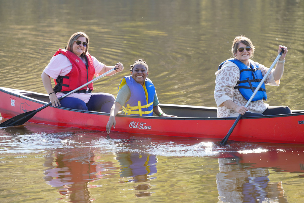 A child smiles while sitting in the middle of a canoe being paddled by two women.