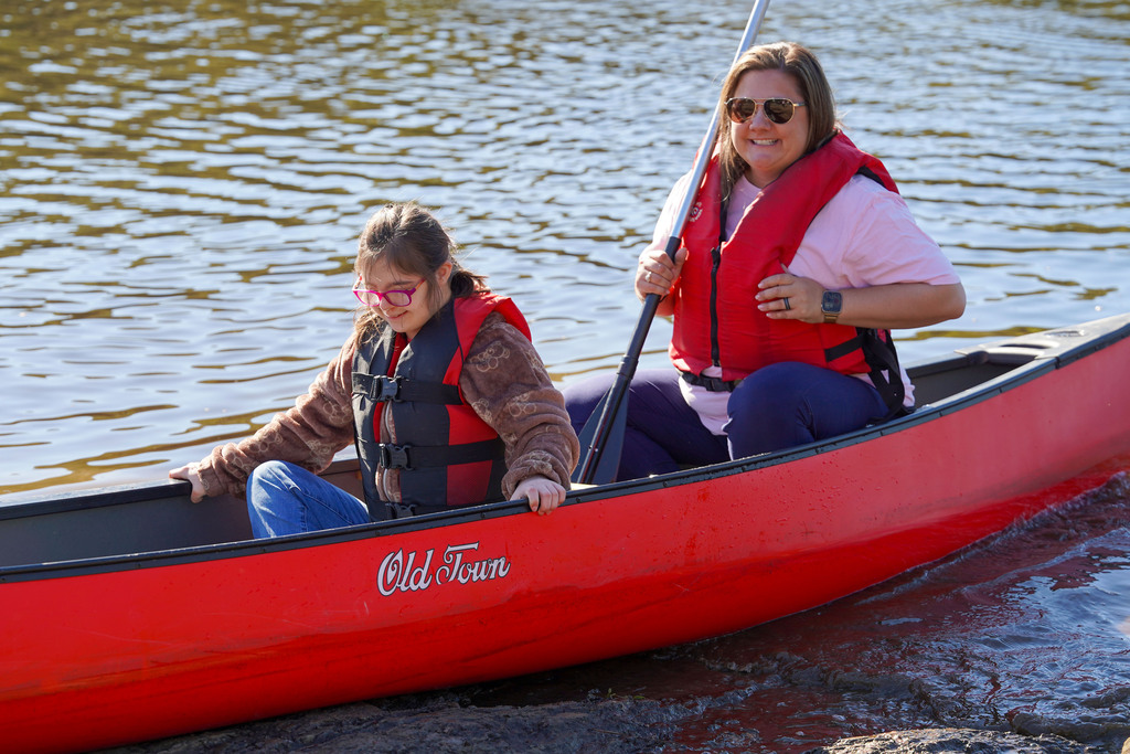 A student smiles while sitting in a canoe on a lake. A woman sits behind her and smiles while holding a paddle.