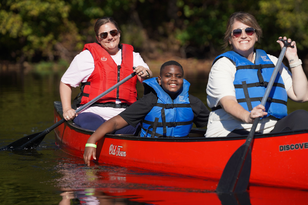 A student smiles while sitting in the middle of a canoe being paddled by two women.