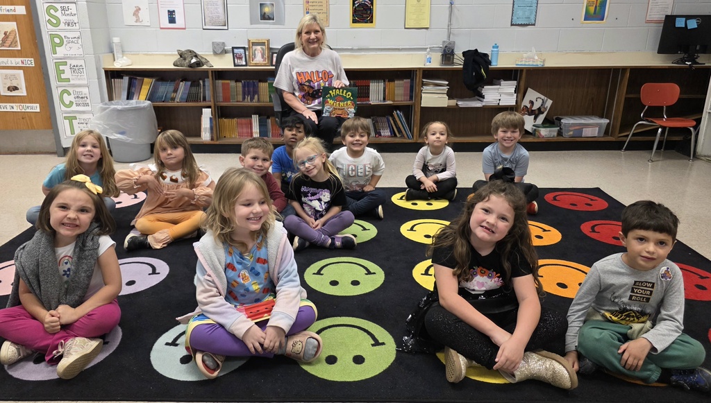 Teacher holding a book and students sitting in front of her on the rug