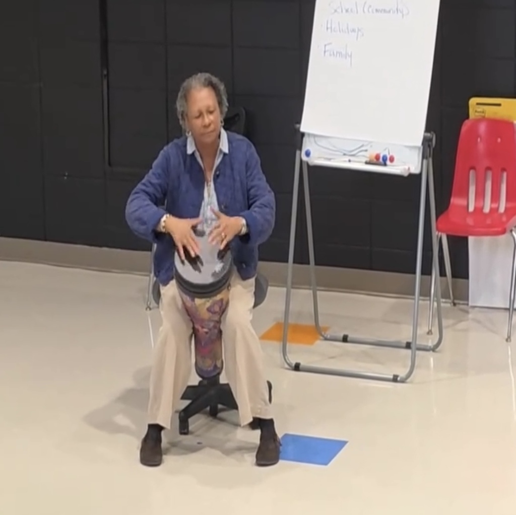 A person sits in a chair in a classroom and plays a drum.